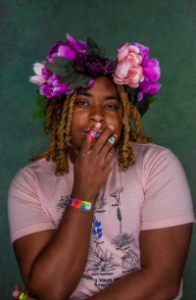 KB — a Black person with brown skin — has a green background. They are wearing a flower crown, with pink and purple flowers, along with green leaves. Their hair is braided and blond. Their hand is slightly covering their mouth, and both their arms and hands have band-aids on them. KB is wearing a pink shirt with black and blue images of cactus plants on them. The elbow of one arm rests on their other arm. They are looking directly at the camera.